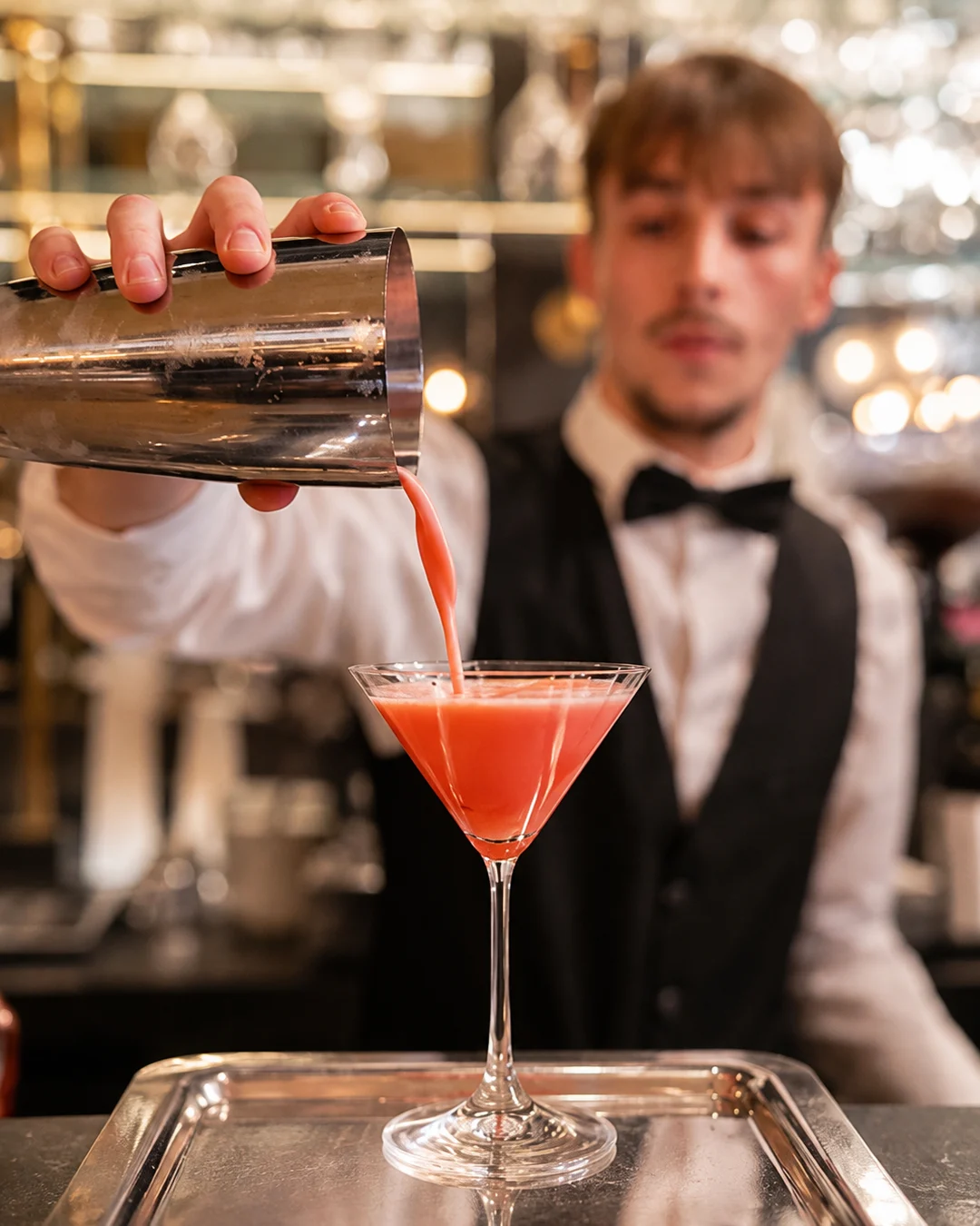 Barman en chemise blanche et nœud papillon préparant un cocktail au shaker derrière un bar dressé en extérieur.